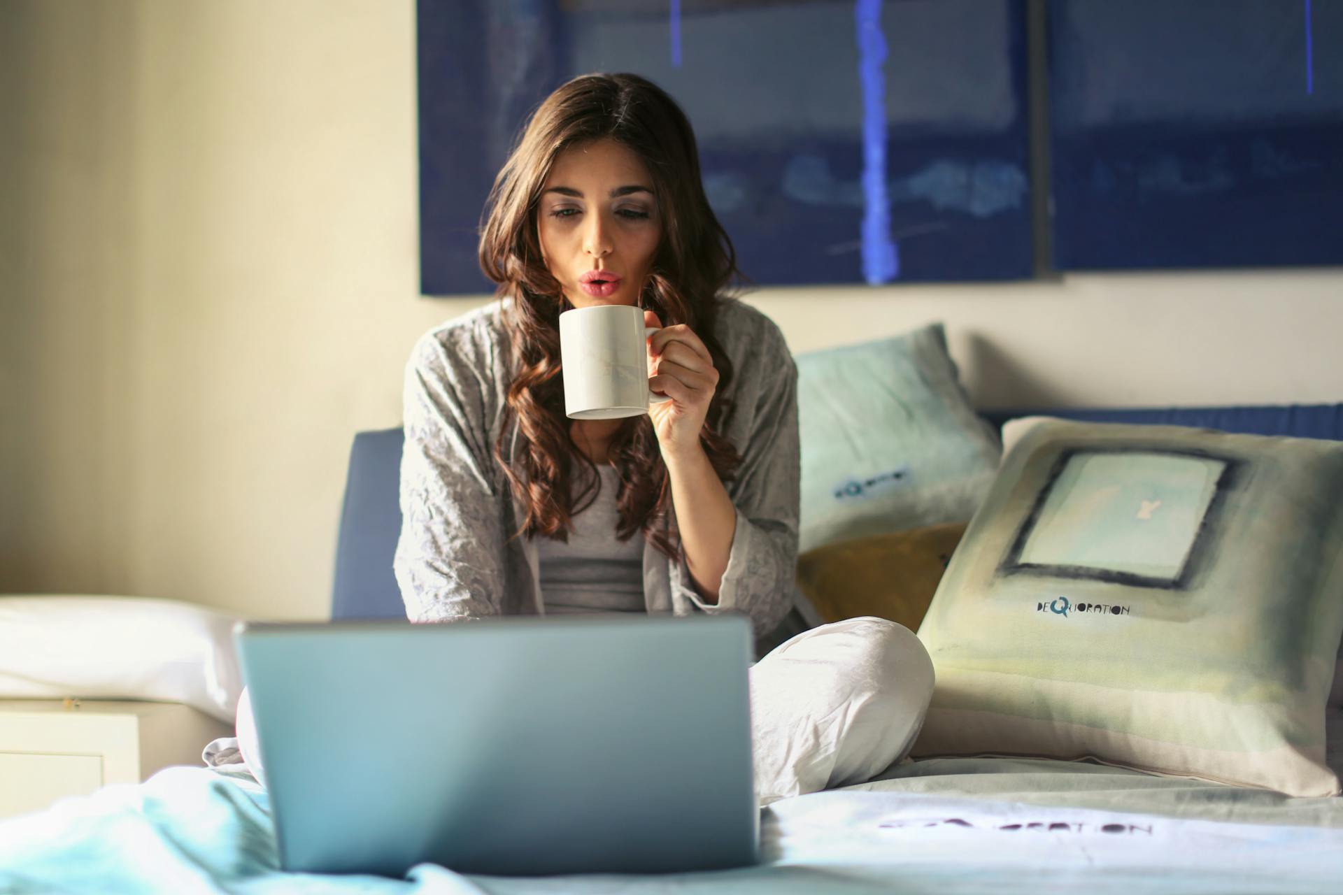 A woman sat on bed, with a hot drink using her laptop
